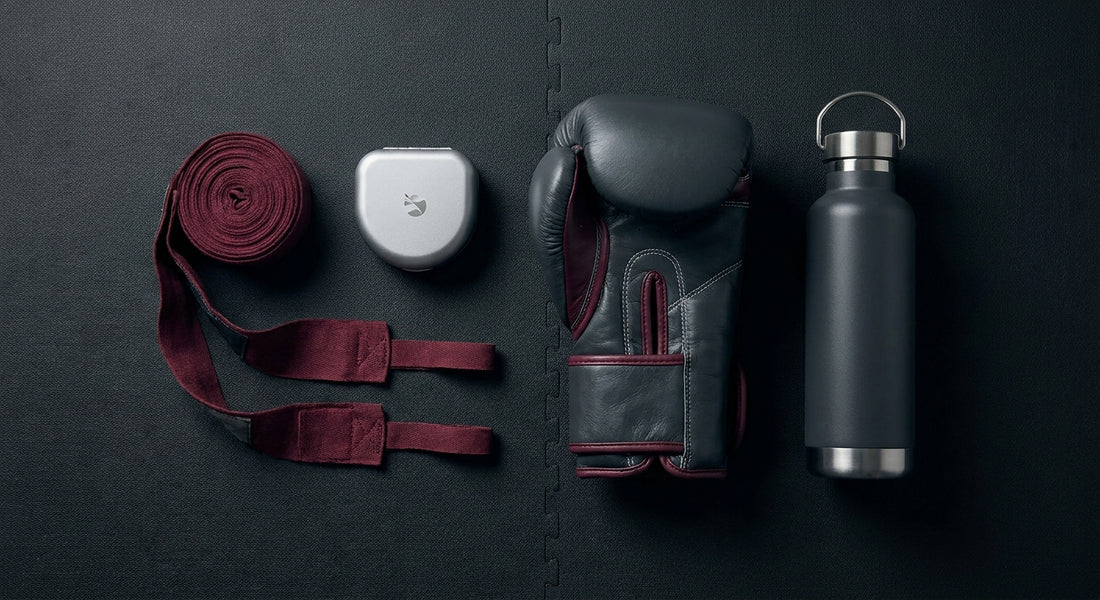 A neatly arranged flat-lay of martial arts training gear including gloves, hand wraps, and accessories on a dark gym mat, styled as a gift selection.