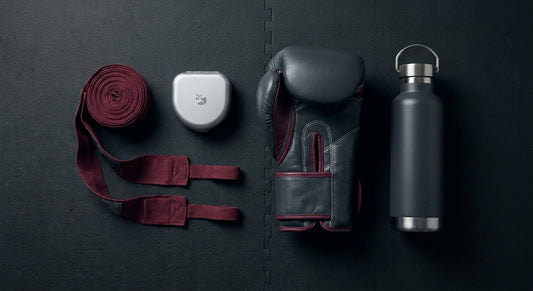 A neatly arranged flat-lay of martial arts training gear including gloves, hand wraps, and accessories on a dark gym mat, styled as a gift selection.