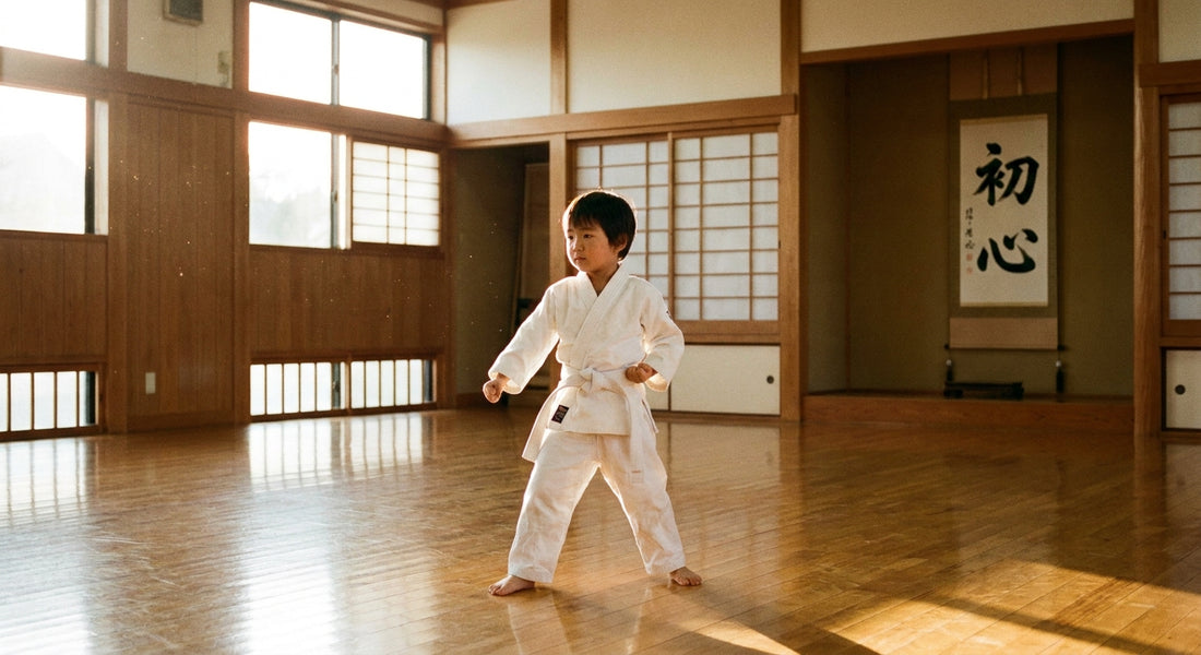 A young child wearing a white martial arts gi stands in a focused ready stance on a wooden dojo floor, bathed in soft natural light.