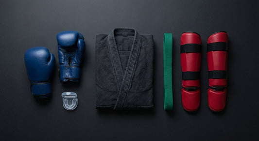An overhead flat-lay of diverse martial arts equipment including boxing gloves, a gi, a belt, shin guards, and a mouthguard arranged on a dark matte surface.